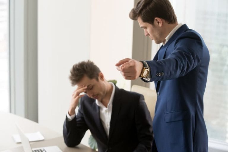 Employee who is suffering workplace retaliation. The boss is pointing for him to leave and the employee is with his head in his hands and looks distressed.