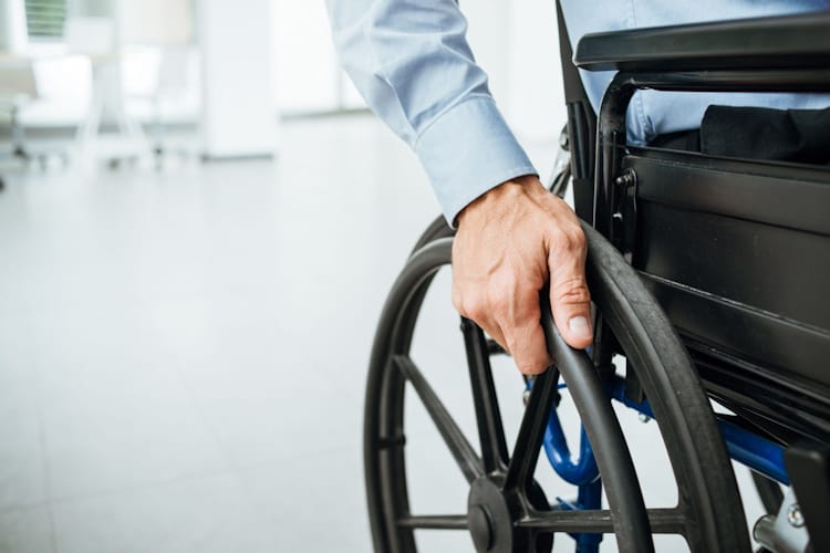 Person's partial body sitting in a wheel chair with the hand holding on to a wheel chair wheel.