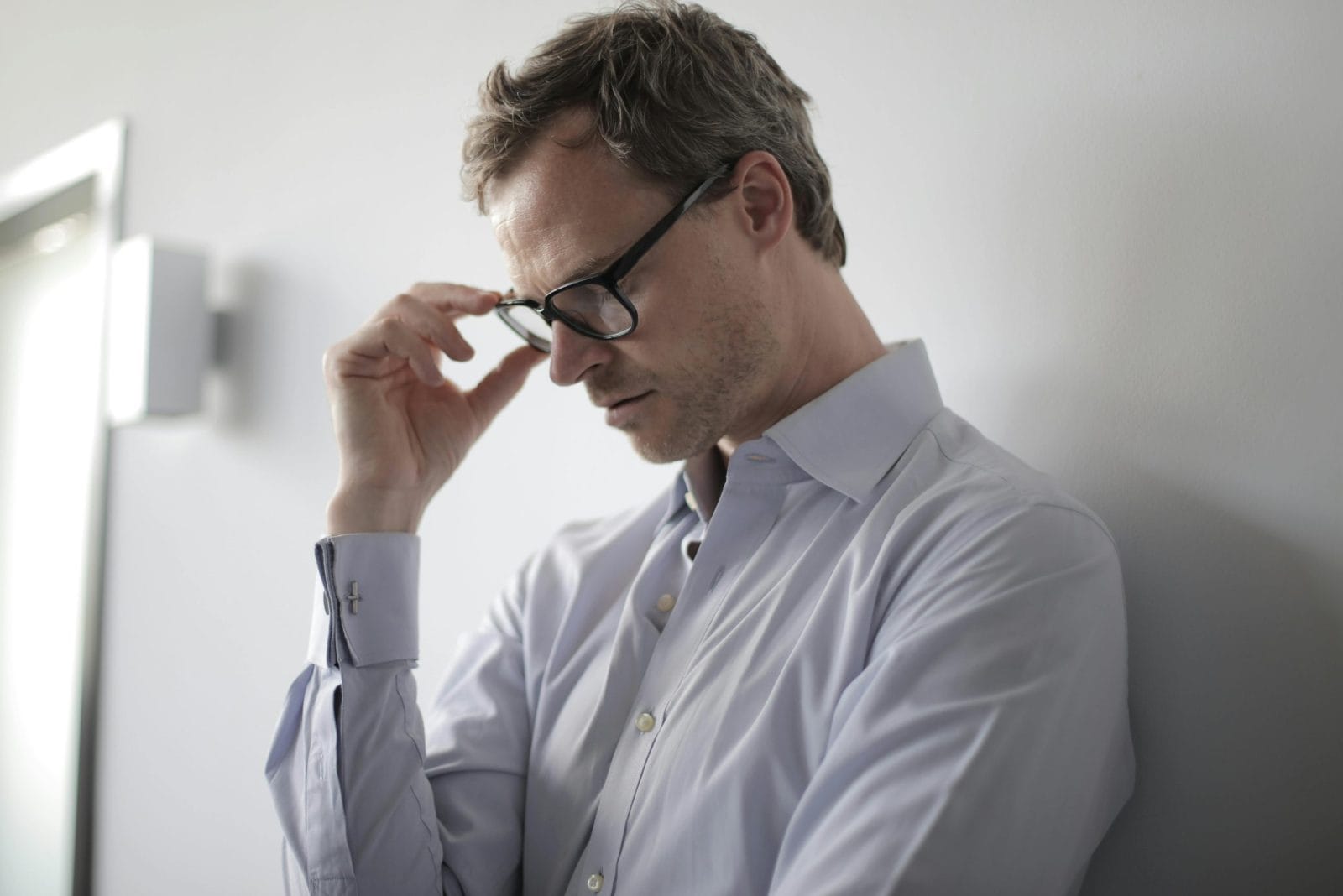 An adult man in a shirt and glasses leaning against a wall indoors he appears stressed due to a workplace retaliation case.