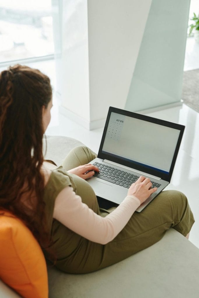 Pregnant woman working from home, sitting on a couch, using a laptop, illustrating remote work and motherhood.