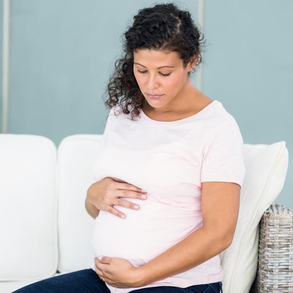 Pregnant lady in a white shirt sitting on a sofa holding her stomach and looking down and sad. 