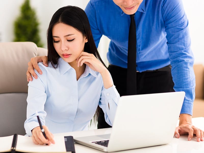 Boss in corporate attire making an unwanted advance on an employee sitting at a desk in front of a laptop. 