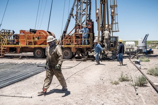 "Workers in safety gear on an active oil drilling site with heavy machinery and pipes on the ground."
The image depicts an outdoor scene at an oil field, possibly in the Permian Basin region.
Foreground: A worker in coveralls, hard hat, and safety glasses is walking across a dusty site.
Midground: Several other workers are present on or near a large orange drilling rig truck.
Background: The area appears arid or desert-like with sparse vegetation. Various pipes and equipment are scattered around the site.