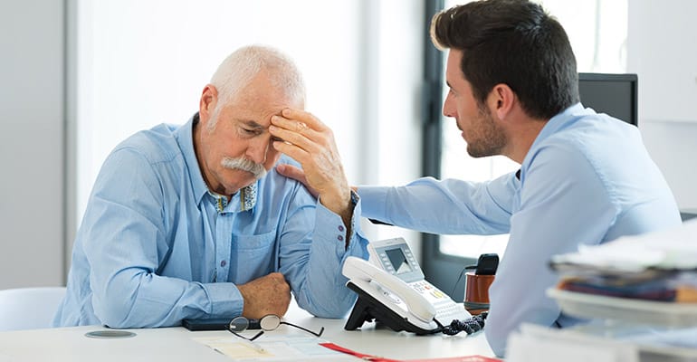 Distressed senior with his head in his hand, and an employer calming him down. They are sitting at a desk with a white office phone.
