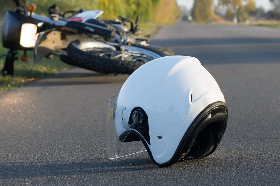 A white motorcycle helmet lies on an asphalt road in the foreground, with a crashed and overturned motorcycle visible in the blurred background, suggesting a recent accident.