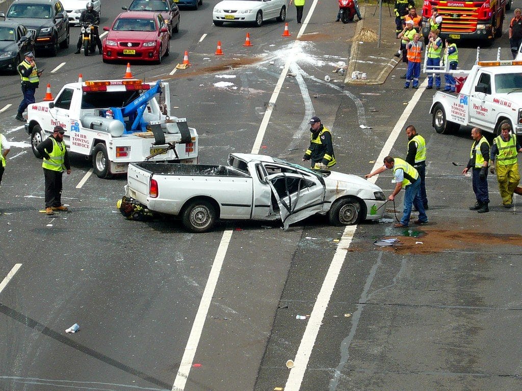 A highway traffic accident scene involving several vehicles and emergency personnel. The image shows a white pickup truck that has sustained significant damage, with its side crushed in. Emergency workers in high-visibility vests and jackets are present, along with two tow trucks. Other vehicles are stopped on the road around the incident.