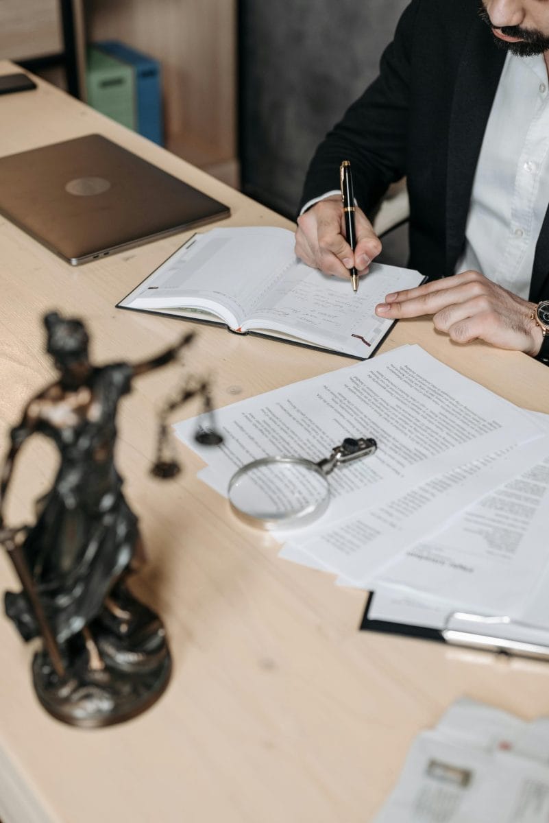 A person in a black suit is writing on a document at a desk, with a Lady Justice statue and a magnifying glass nearby. The scene suggests a legal or business setting where documents are being reviewed or signed.