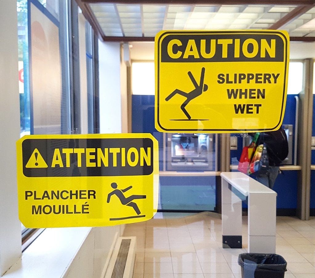 Two yellow caution signs are displayed on a glass door inside a building. The top sign, in English, reads CAUTION SLIPPERY WHEN WET and includes a graphic of a figure slipping. The bottom sign, in French, reads ATTENTION PLANCHER MOUILLÉ and features a similar slipping figure graphic."
