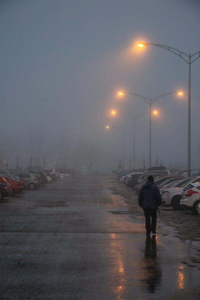 A person walks through a foggy, wet parking lot illuminated by warm streetlights.