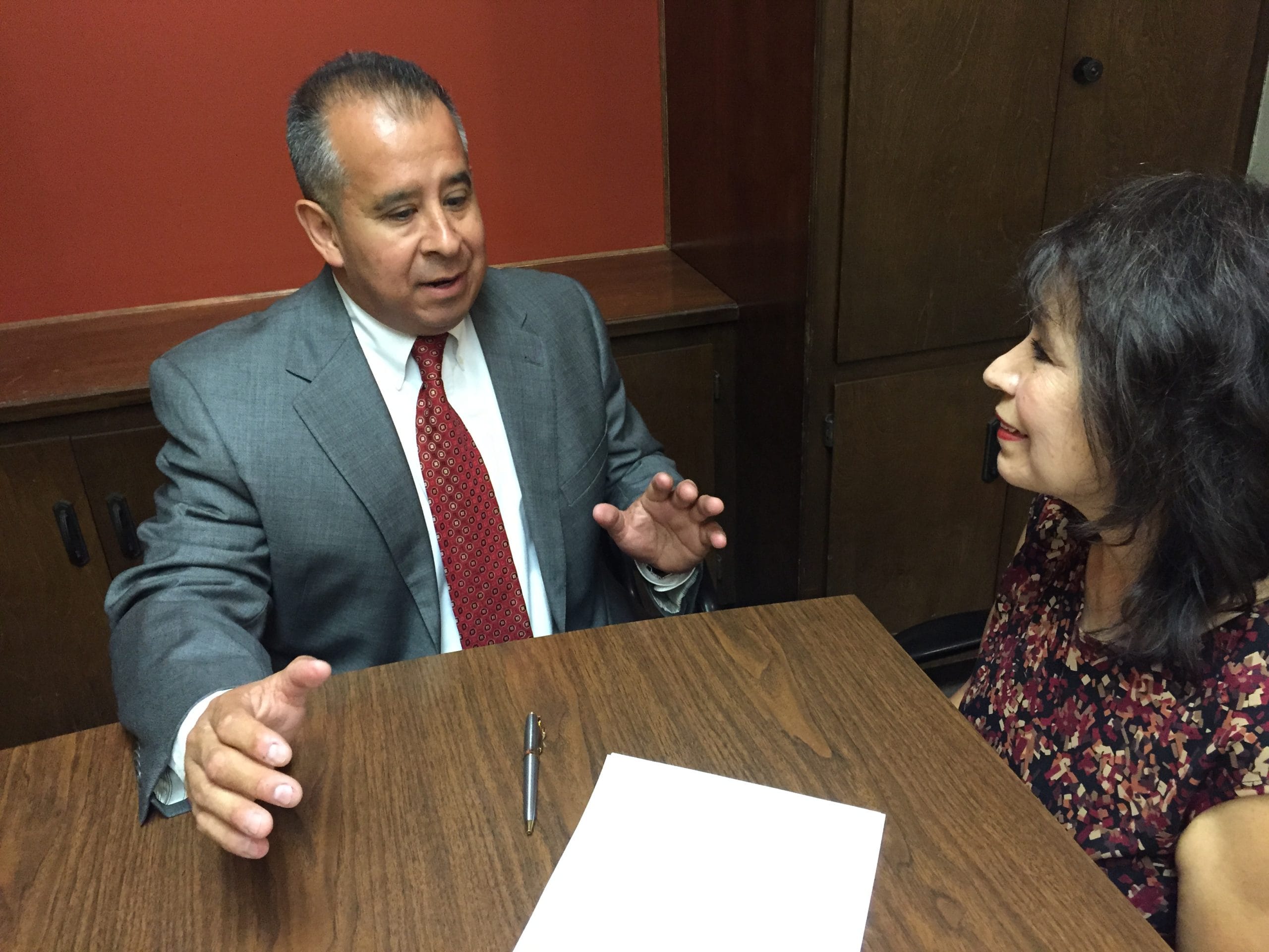 This image shows two individuals having a conversation seated at a table.
One individual is wearing a suit and tie while gesturing with their hands.
The other individual is listening attentively.
They are in an office setting.
A pen and a blank sheet of paper are on the table in front of them.